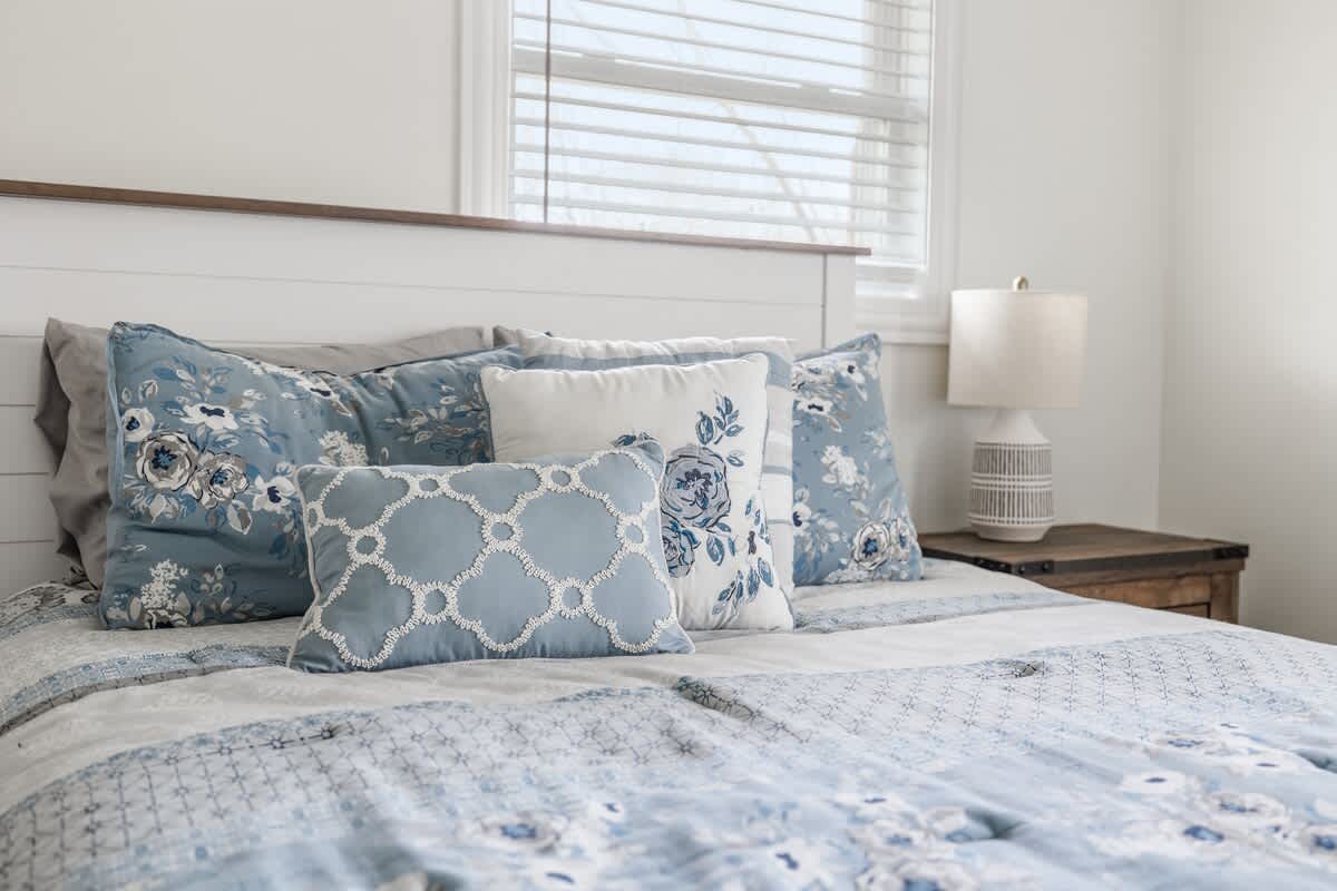 Close-up of the bed in bedroom-1, showing blue and white patterned pillows and bedding, a white and wood headboard, and a white textured lamp on a wooden nightstand.