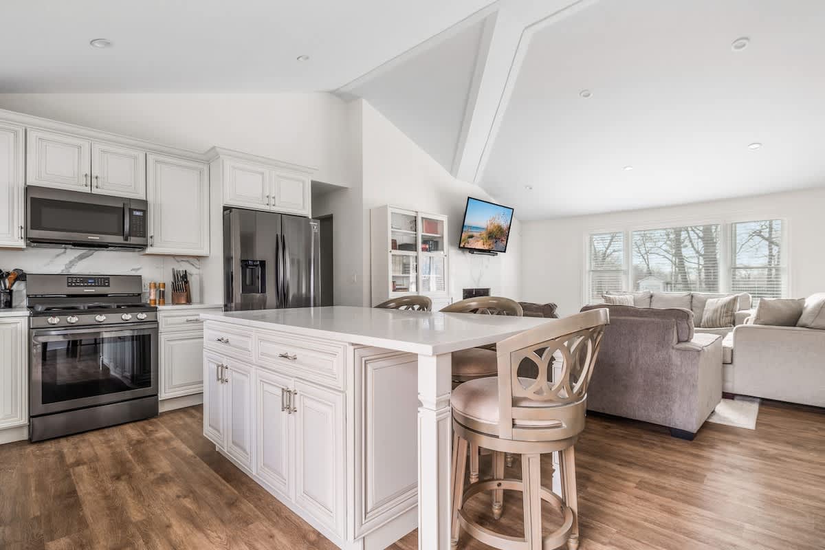 Open concept kitchen and living room with a white kitchen island, white cabinets, stainless steel appliances, and a gray sectional sofa in the background, under a vaulted ceiling.