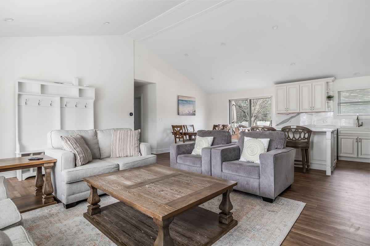 A bright living room with light gray sofas and a large wooden coffee table opens to a dining area and kitchen in the background. A white coat rack and storage unit are visible near the entrance, and large windows offer views of the outdoors.