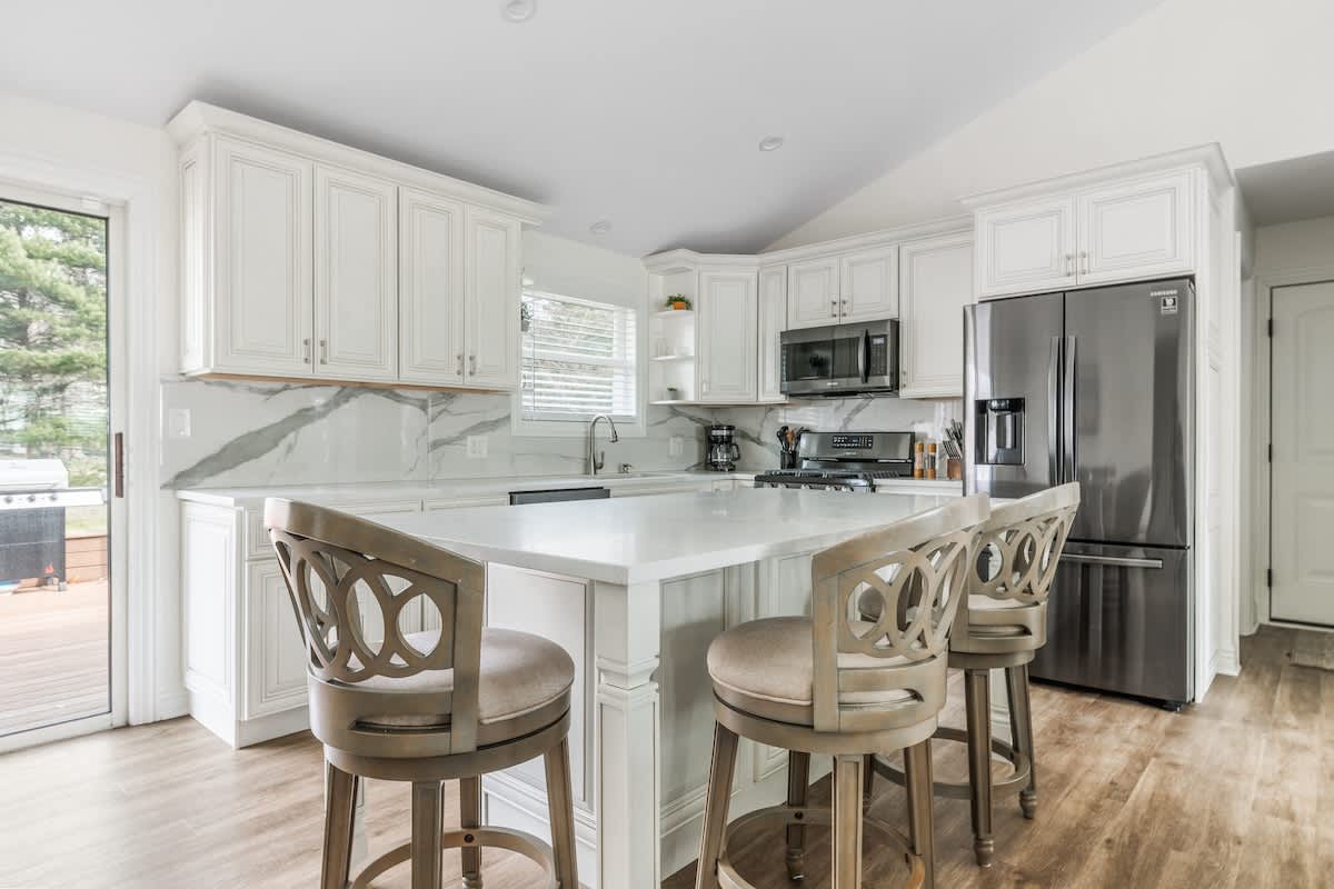 Wide view of a bright kitchen with white shaker cabinets, a large white island with seating, stainless steel refrigerator and stove, and a sliding glass door leading to an outdoor deck.
