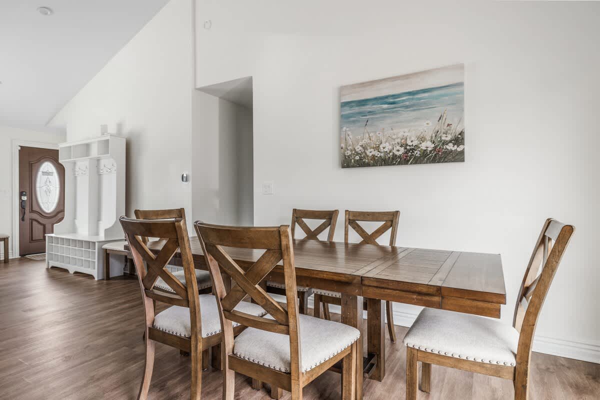 Wooden dining table with six chairs in an open dining area, featuring wood-look flooring, white walls, and a coastal-themed painting, with a mudroom bench visible in the background.