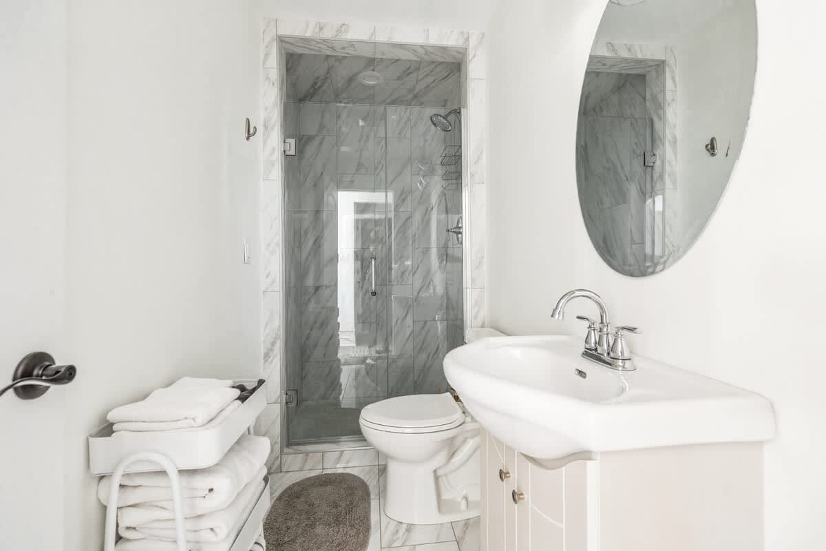 Modern bathroom with a white pedestal sink, a large oval mirror, and a walk-in shower with gray marble-look tile and a glass door. White walls and floor tiles complete the bright space.