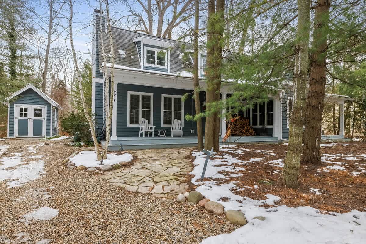 The charming blue-sided house with white trim features a covered front porch and a stone pathway leading to the entrance. A small matching shed is visible to the left, all surrounded by snow-dusted ground and tall trees.