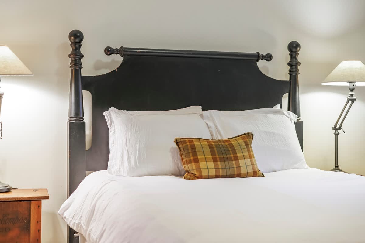 A cozy bedroom features a black wooden four-poster bed with white bedding and a plaid throw pillow, flanked by two table lamps on wooden nightstands.