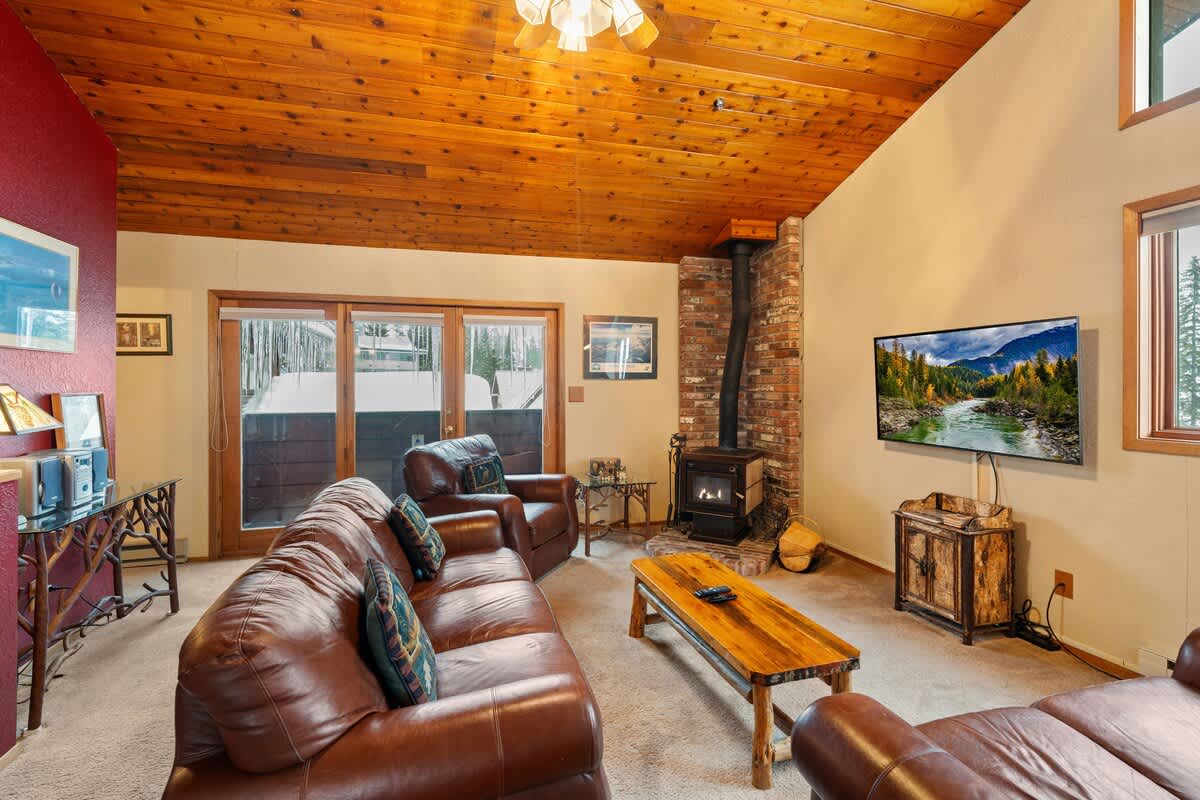 Warm living room with a wood-paneled ceiling and light-colored carpet, featuring a brick-backed wood-burning stove, a long brown leather sofa, and a sliding glass door leading to an outdoor area.