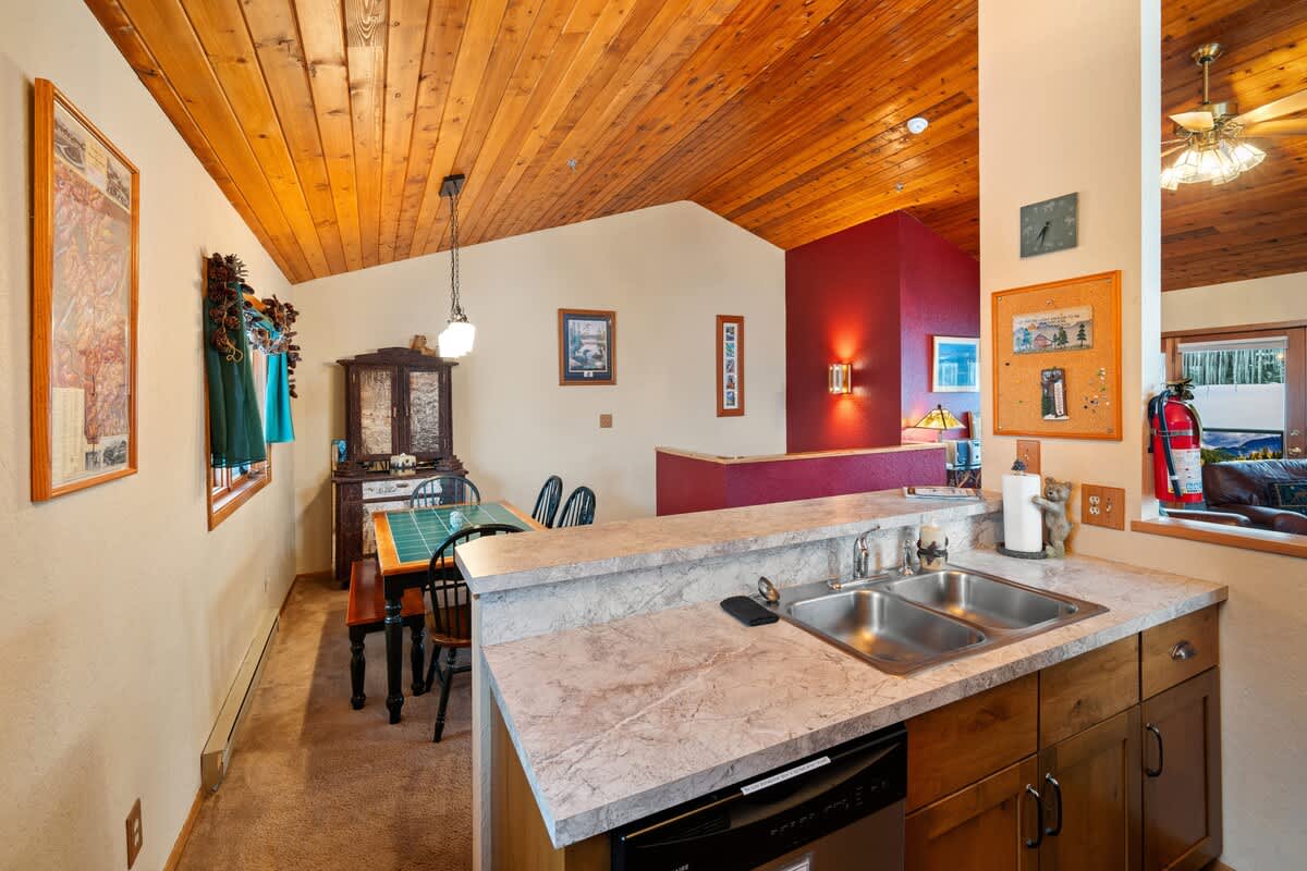 Kitchen area featuring a double sink set in a light-colored countertop, a dishwasher, and wood cabinetry, with a view towards the dining area and a red accent wall.