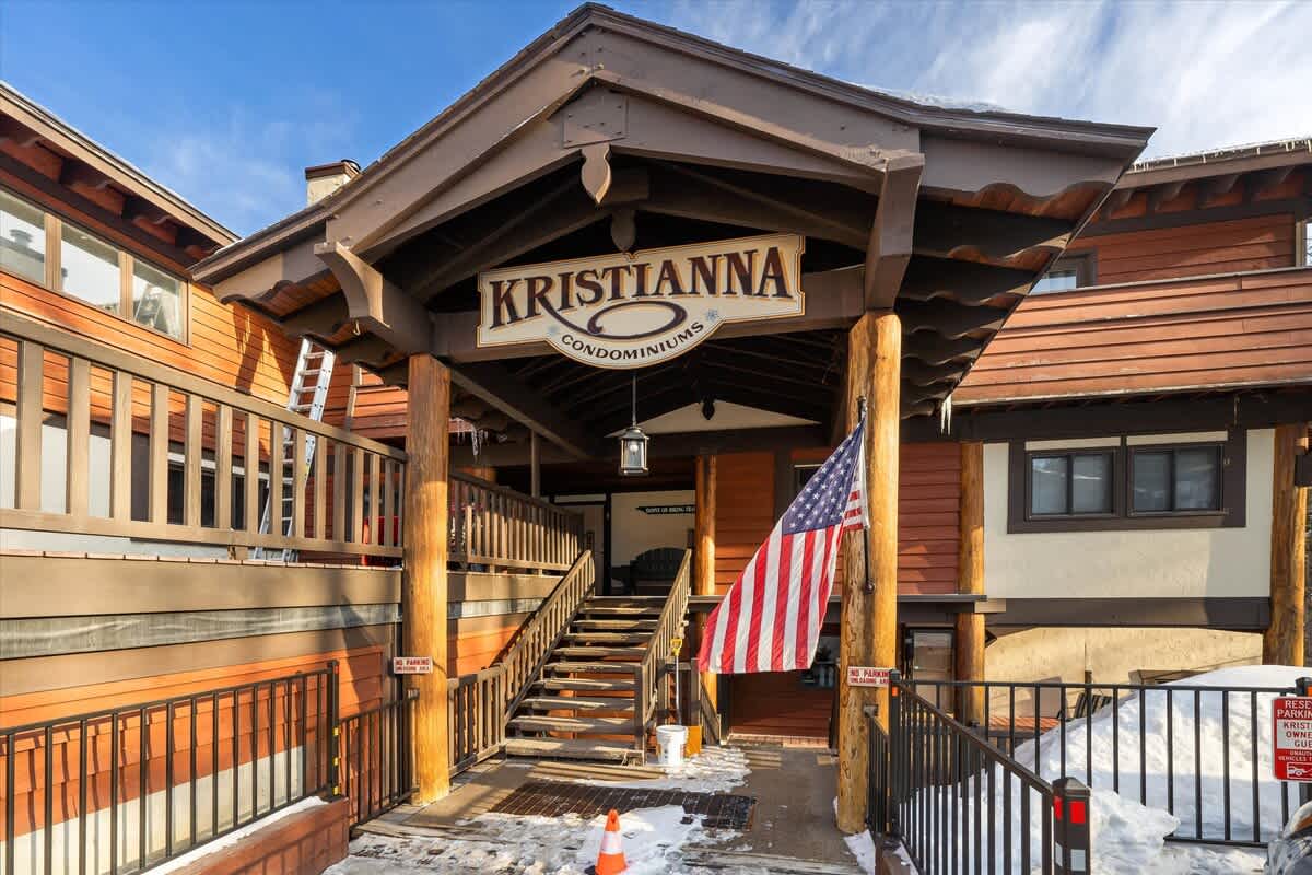 The main entrance of the "Kristianna Condominiums" building, featuring a large wooden sign, a covered porch, and a set of wooden stairs. An American flag hangs near the entrance, and snow is visible on the ground and roof.