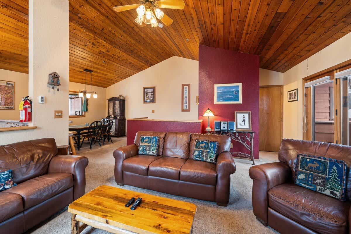 Inviting living room with a vaulted wood-paneled ceiling, furnished with brown leather seating and a wooden coffee table, with a partial view into the dining area beyond a low wall.