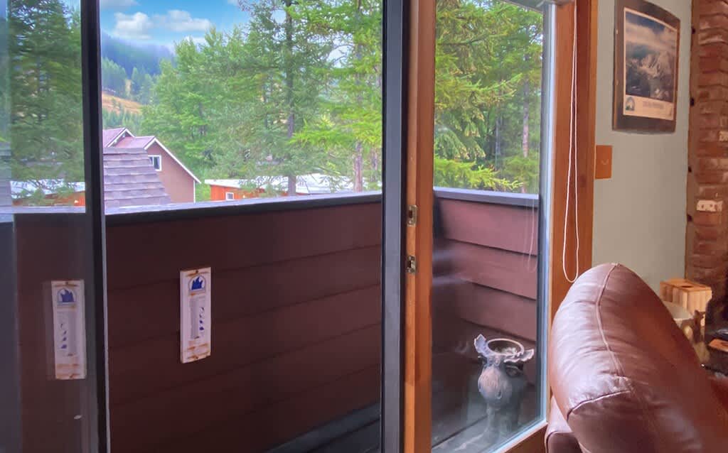 A view through a glass sliding door onto a small balcony, revealing a lush green mountainous landscape with other houses in the distance, suggesting an outdoor space with scenic views.