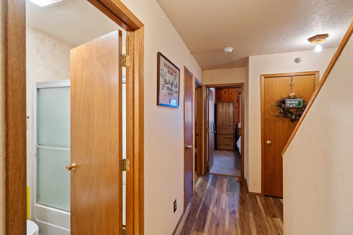 A hallway with light wood flooring and neutral walls, featuring several wooden doors, one of which leads into a bathroom with a shower. A framed picture hangs on the wall, and a staircase is visible on the right.