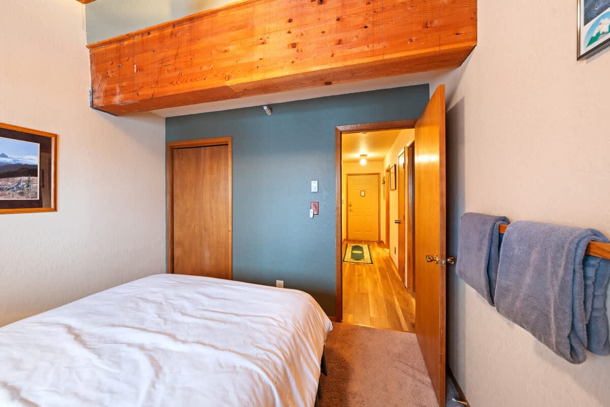 Another view of bedroom 2, showing the queen bed with white bedding, a wooden beamed ceiling, and a doorway leading to a hallway with wooden flooring, emphasizing the room's layout and connection to the rest of the property.