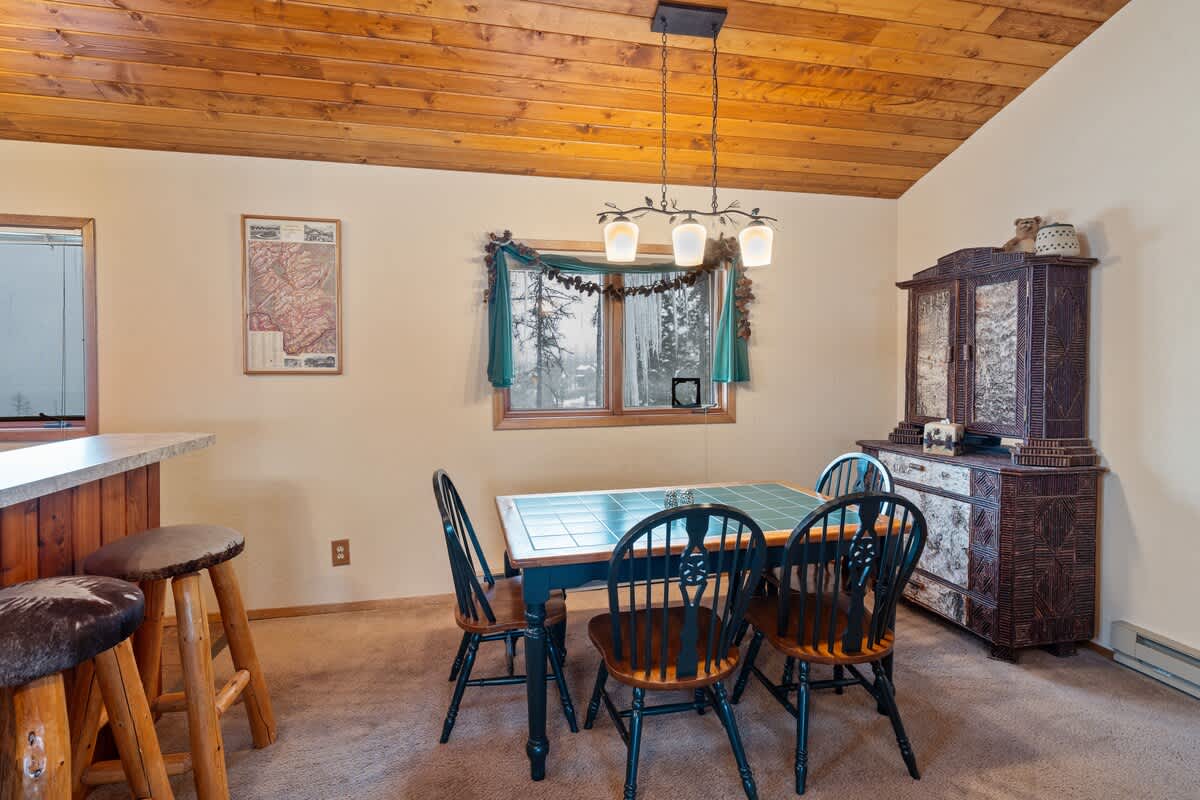 A cozy dining area with a wooden table and four black spindle-back chairs sits on a carpeted floor, illuminated by a rustic chandelier, with a decorative cabinet and a window adorned with winter garland.