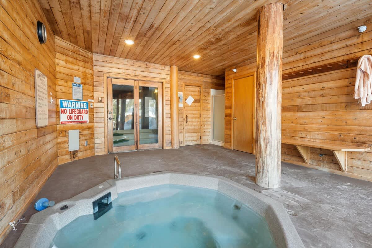 An indoor hot tub area with rustic wood-paneled walls and ceiling, featuring a large grey hot tub with steps. A "WARNING NO LIFEGUARD ON DUTY" sign is prominently displayed, and there's a wooden bench and a glass door leading outside.