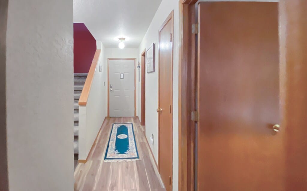 A long, narrow foyer with light wood flooring and a blue patterned runner rug leading towards a white front door. A staircase with a wooden handrail is visible on the left, and walls are painted in a neutral tone with a dark red accent wall.