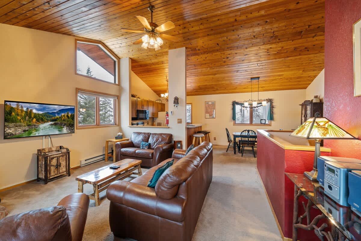 Open-concept living space with a vaulted wood-paneled ceiling, featuring a living area with leather sofas, a breakfast bar separating it from the kitchen, and a dining area visible in the background.