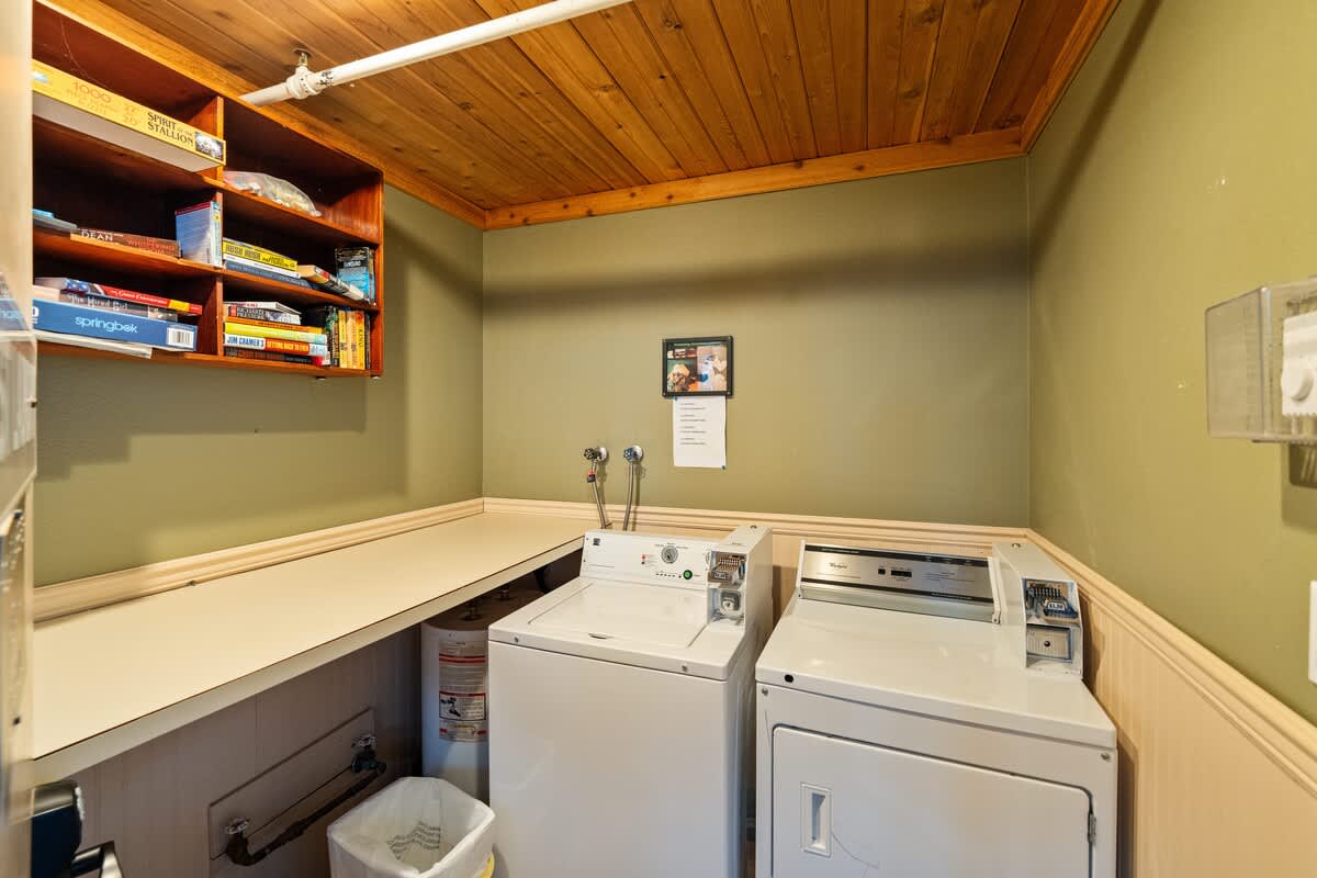 A dedicated laundry room with a white top-loading washing machine and a front-loading dryer, set beneath a light-colored countertop. A wooden shelf filled with board games is mounted above, and the room has olive green walls and a wood-paneled ceiling.