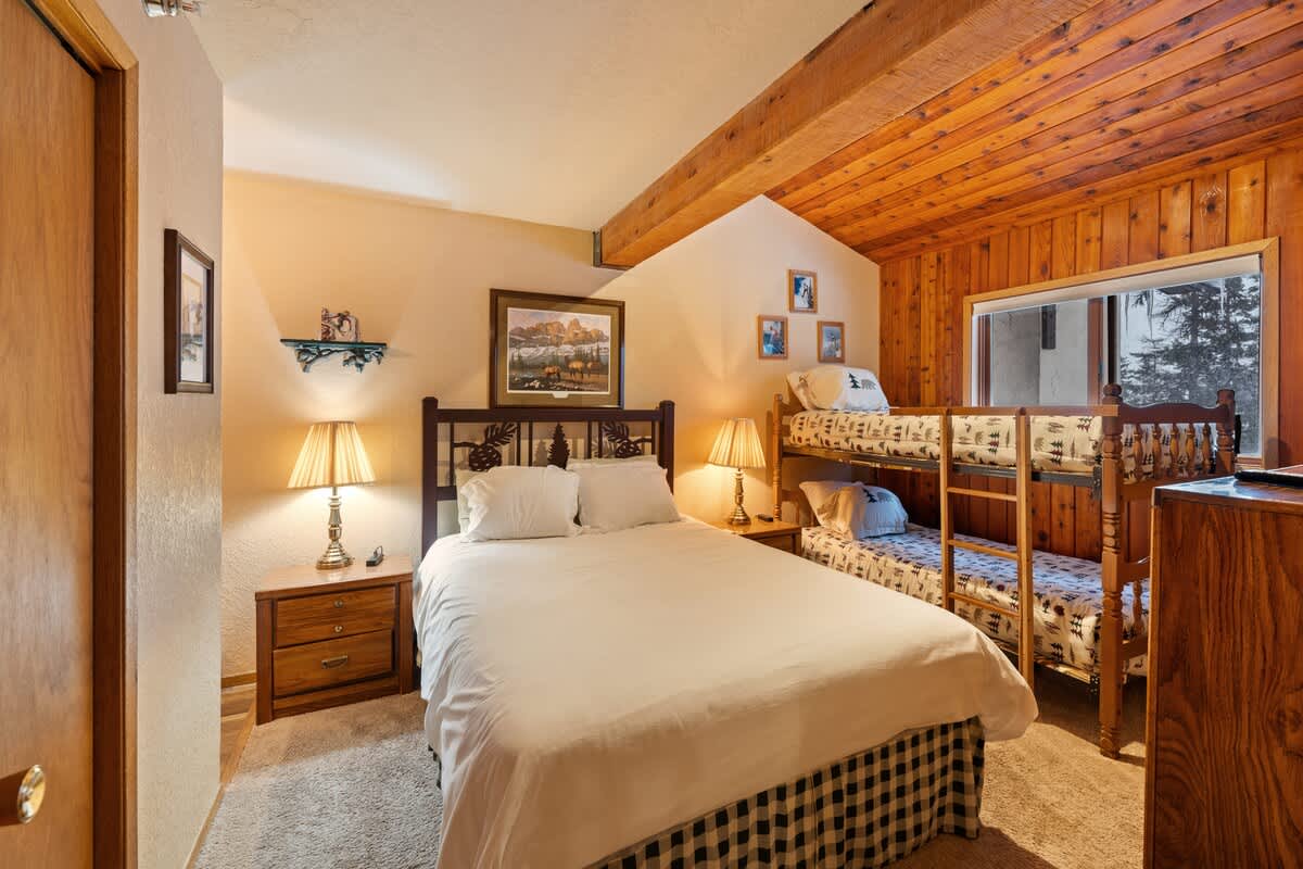 A wide shot of bedroom 2 showcasing a queen bed with a dark wooden headboard and a bunk bed, both with white bedding, under a wooden ceiling, with bedside lamps and framed artwork adding to the cozy ambiance.