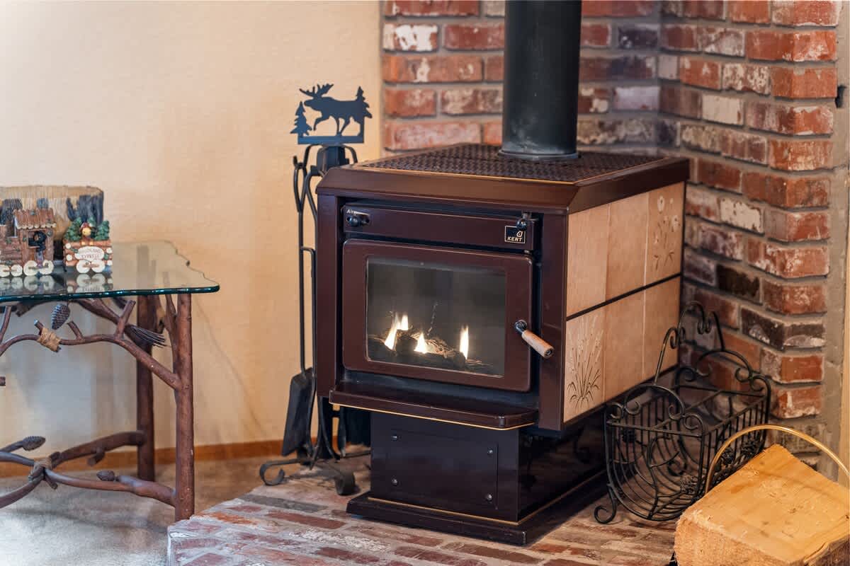 Close-up of a rustic wood-burning stove with a glass door, set on a brick hearth against a brick wall, with a moose-themed fire tool set nearby.