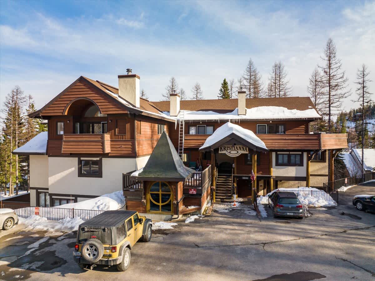 An aerial view of the "Kristianna Condominiums" building, a multi-story structure with brown siding and a gabled roof, surrounded by snow-covered ground and pine trees. Several cars are parked in the foreground, and the entrance is visible.