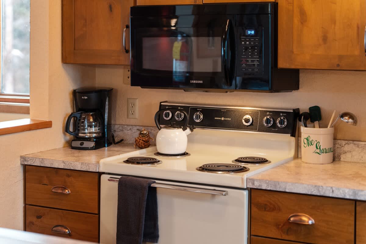 A rustic kitchen features wooden cabinetry, a black microwave above a white electric stove, and a coffee maker on the countertop, creating a functional and cozy cooking space.