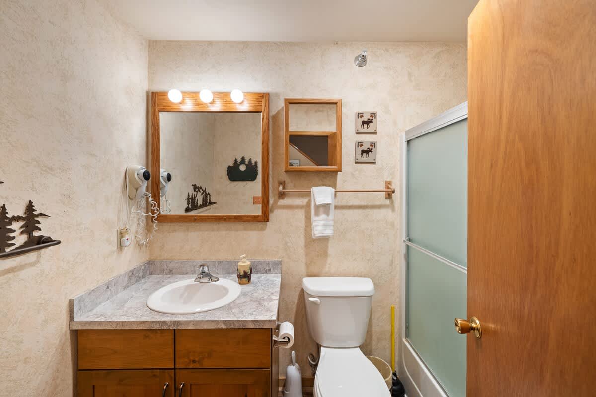 A rustic-themed bathroom featuring a wooden-framed mirror with globe lights, a single vanity with a white sink and speckled countertop, and a toilet next to a glass-enclosed shower. The walls have a textured, light-colored finish.