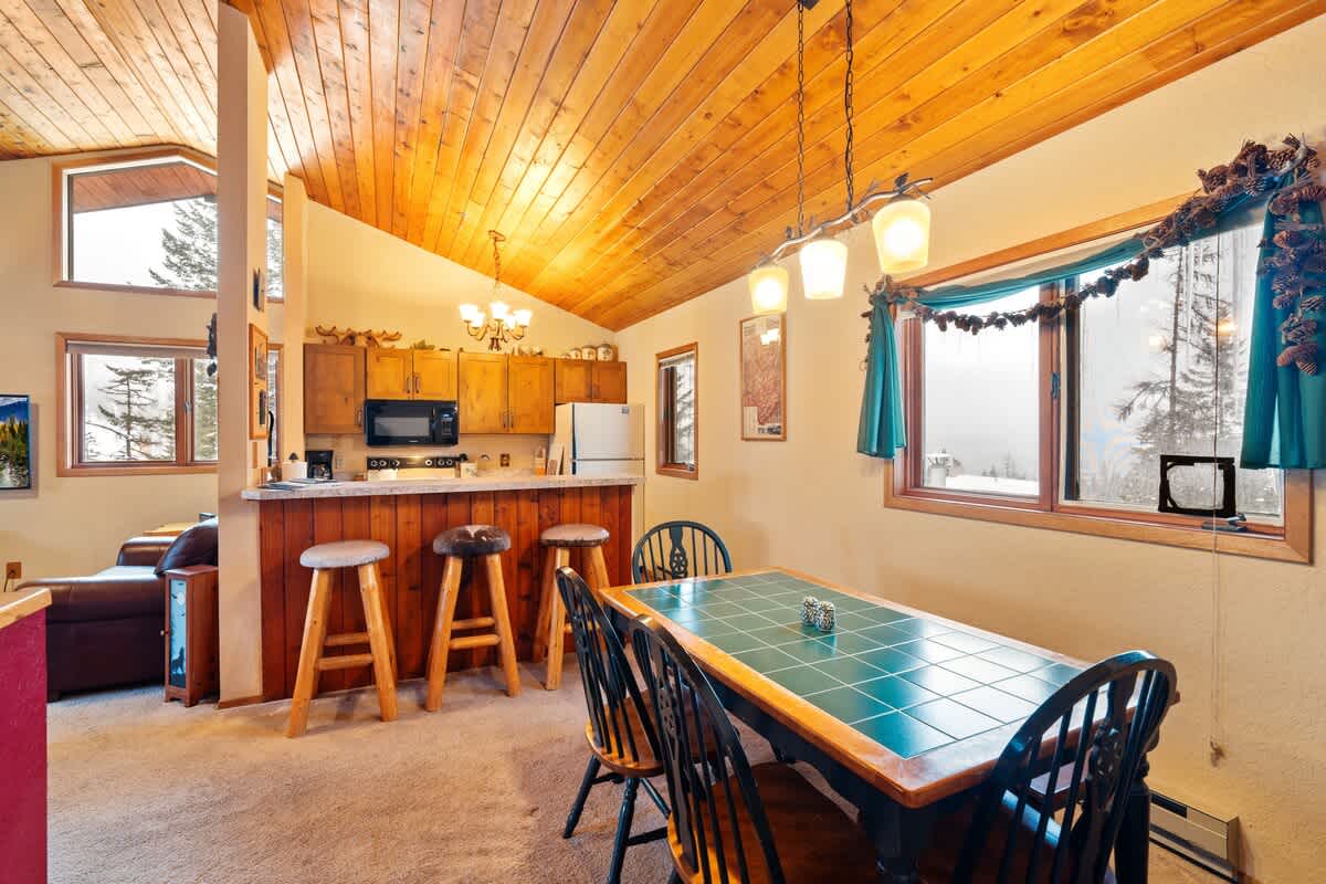 Dining area with a wooden table and chairs, illuminated by a chandelier, adjacent to a breakfast bar with stools, and featuring a wood-paneled ceiling and a window with a festive garland.