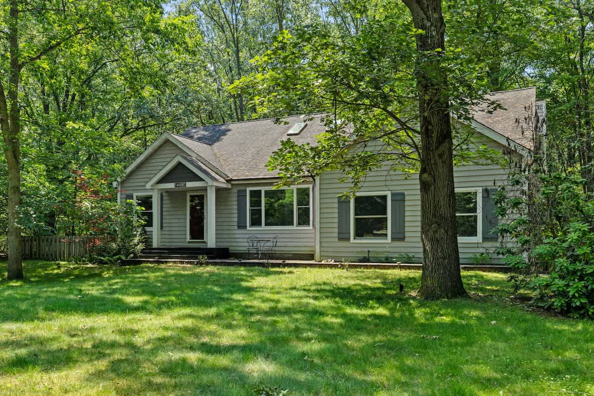 The front exterior of a charming gray-sided house with white trim and dark shutters, nestled among green trees and featuring a small front porch with a dark door. A small table and chairs are visible on the front lawn.