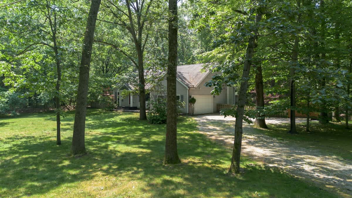 The front exterior of the house, partially obscured by tall trees, shows a light-colored siding and an attached garage, with a gravel driveway leading up to it, surrounded by a lush green lawn.