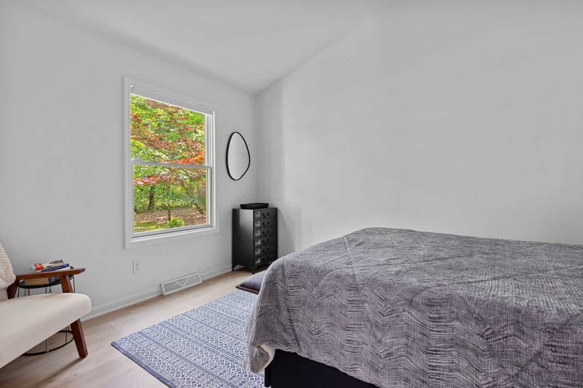 This bright bedroom features a queen-sized bed with gray bedding and a blue patterned rug, with a window offering a view of lush green trees. A black dresser and a small round mirror are visible on the far wall.