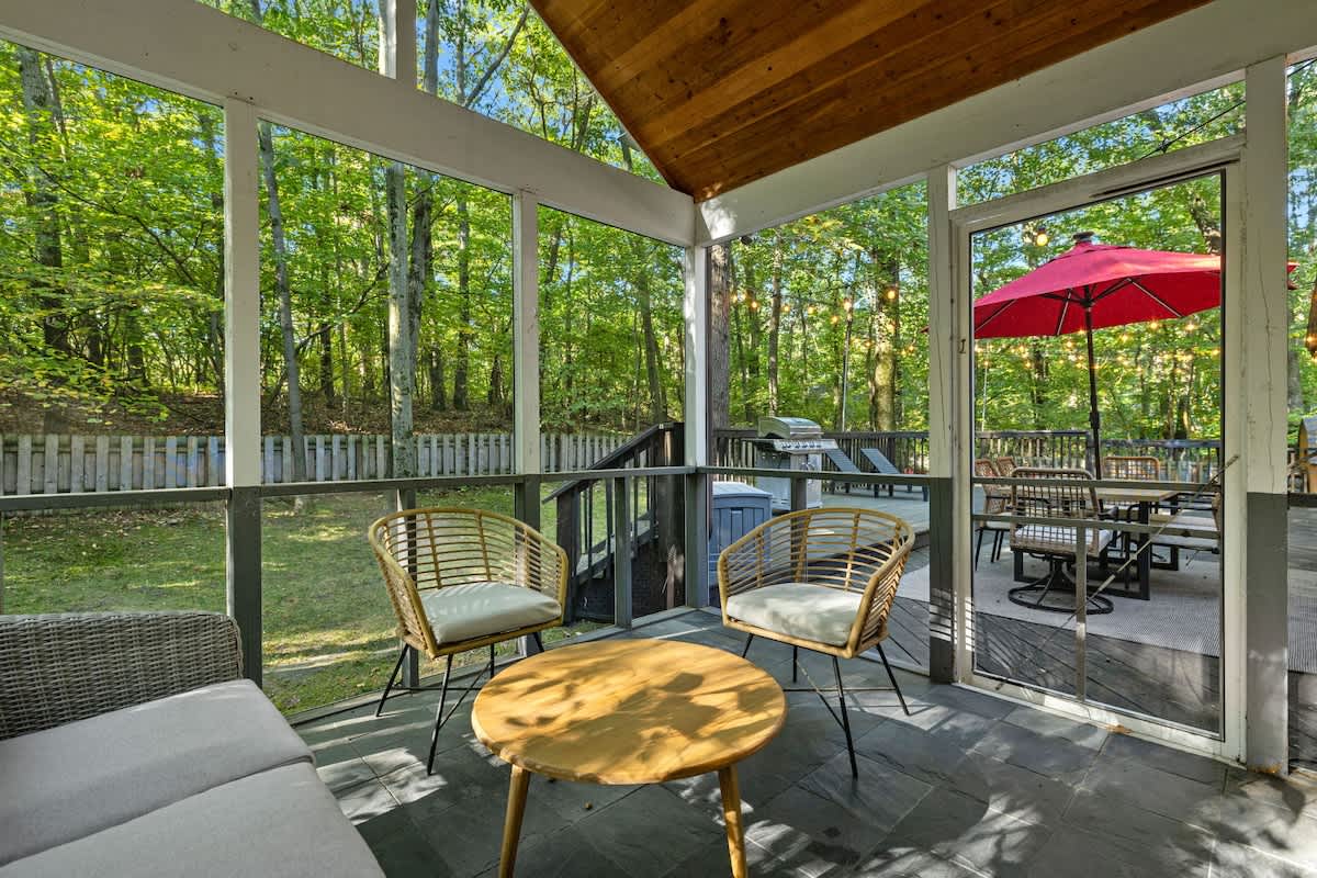 A spacious screened porch with comfortable outdoor seating, including a gray sofa and two wicker chairs around a wooden coffee table. Beyond the screen, a deck with a red umbrella and dining set is visible, surrounded by lush trees.