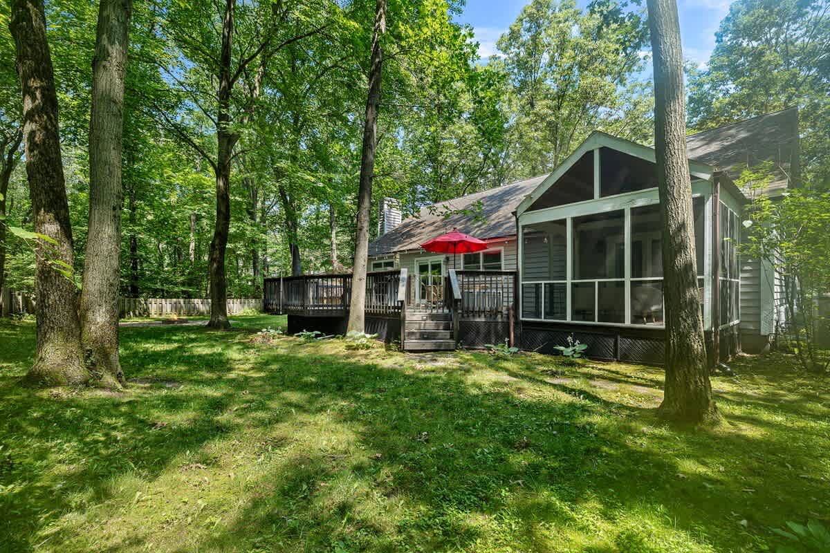 The back of the house is visible from the grassy yard, featuring a multi-level dark wooden deck with stairs leading down, a screened porch, and a red umbrella, all nestled among tall trees.