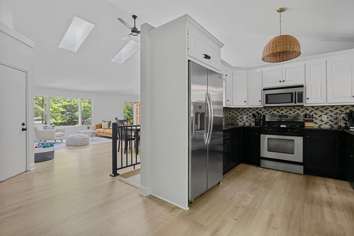 An open-concept view showcasing the kitchen with white and black cabinetry and stainless steel appliances, flowing into a bright living area with light wood floors, vaulted ceilings, and skylights.