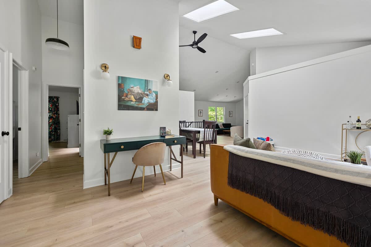 Open-concept living space with vaulted ceilings and skylights, featuring a dark green desk area, a brown leather sofa, and a dining area visible in the background.