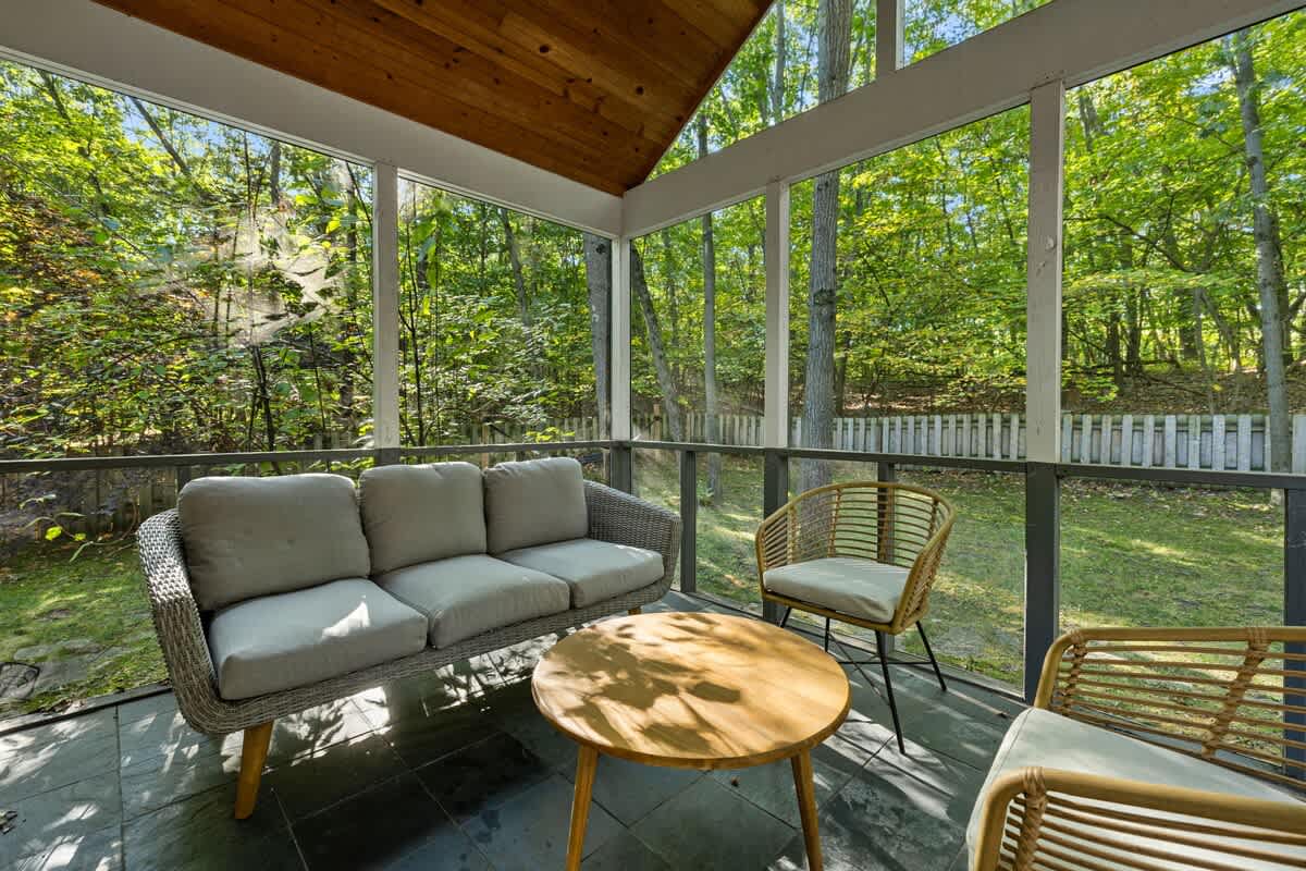 A cozy screened-in porch features a light gray wicker sofa with cushions, a matching armchair, and a round wooden coffee table, all set on dark slate tile flooring with a view of the surrounding woods.