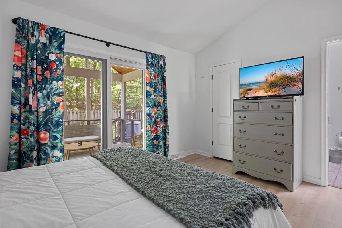 This bedroom offers a view of a large flat-screen TV mounted on a gray dresser, with vibrant floral curtains framing sliding glass doors that lead to a screened porch. A white bed with a green throw blanket is in the foreground.