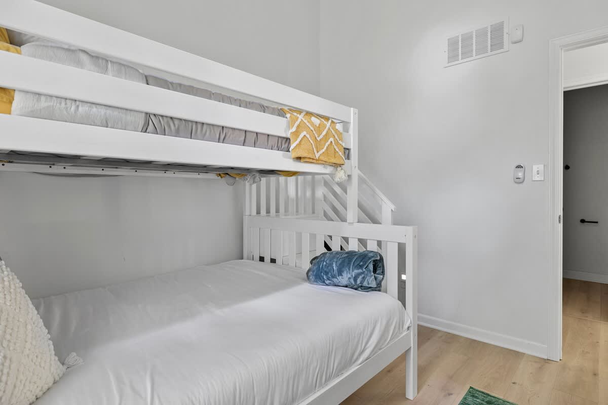 A close-up view of the white wooden bunk bed in a children's room, showing the full-sized bottom bunk with a blue throw and the twin top bunk with yellow and gray bedding. The built-in stairs for the top bunk are visible.