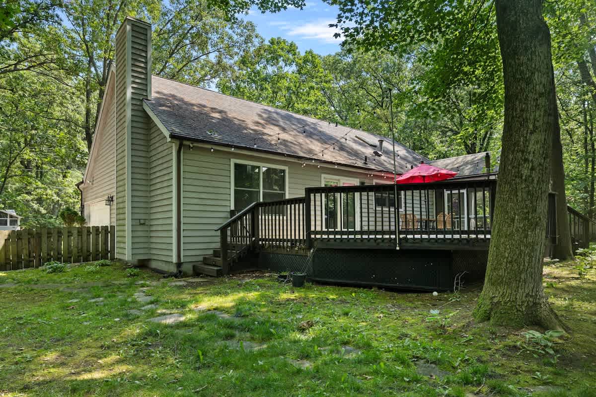 The back of a light-colored house with horizontal siding and a prominent chimney, featuring a multi-level dark wooden deck with a red umbrella and surrounded by lush green trees and grass.