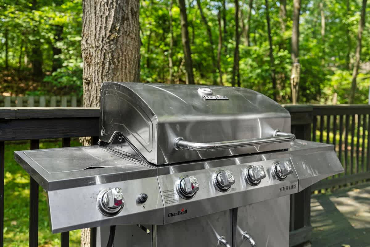 Close-up view of a stainless steel Char-Broil gas grill with four burners and side tables, positioned on a dark wooden deck surrounded by lush green trees.