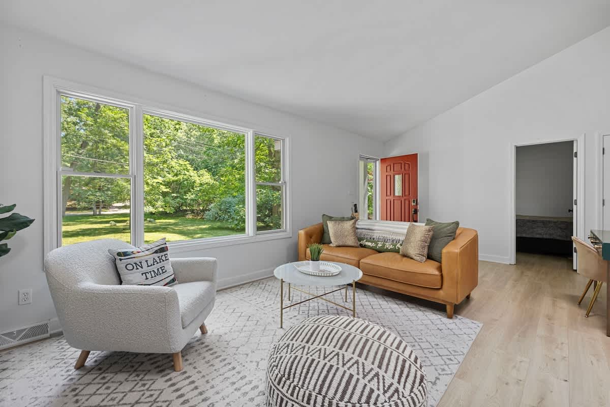 Bright living room with a white armchair, a brown leather sofa, and a round glass coffee table on an area rug, featuring large windows overlooking a lush green outdoor space.