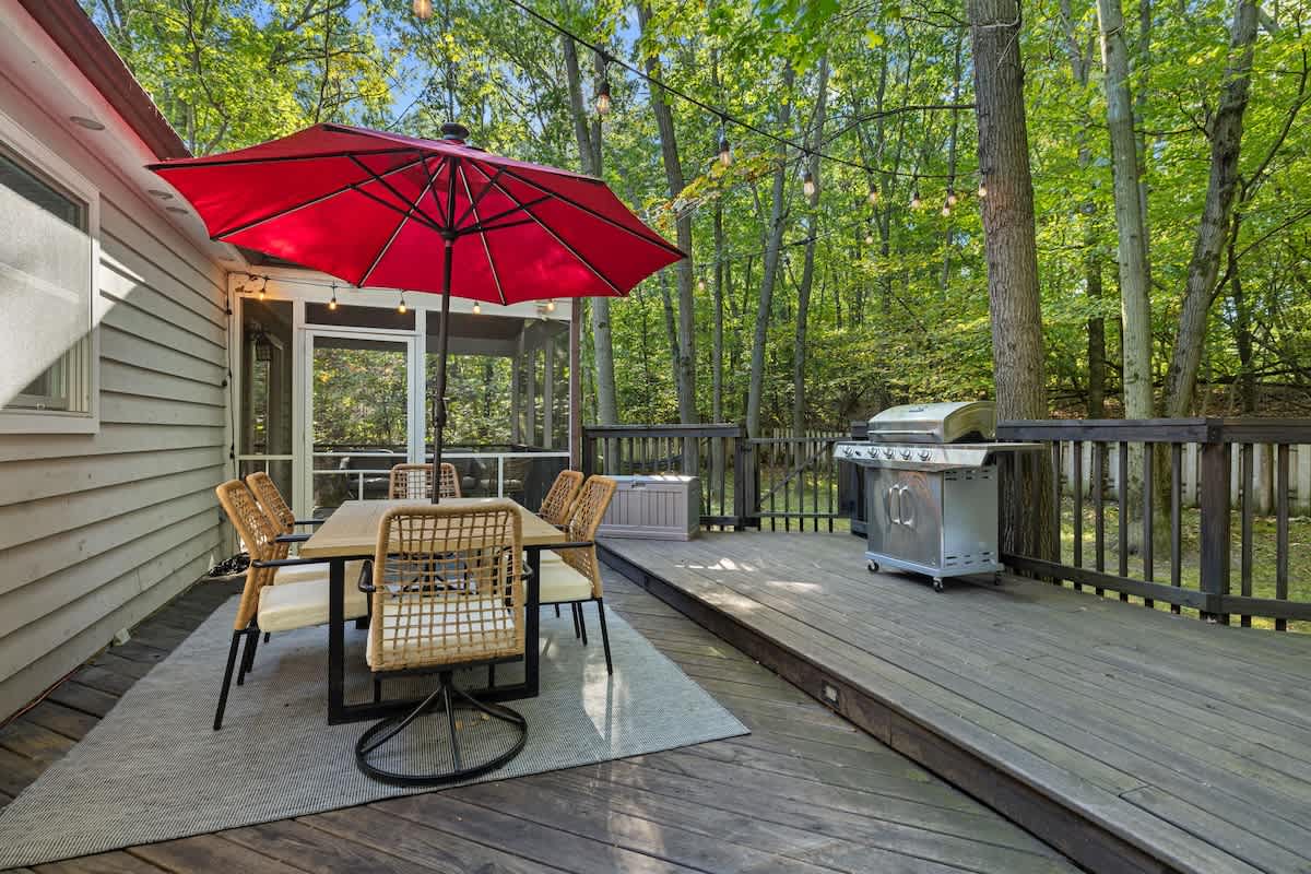 An outdoor dining area on a dark wooden deck, featuring a rectangular table with six wicker chairs under a large red umbrella, with a stainless steel grill nearby and string lights overhead.
