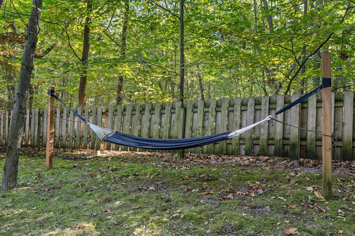 A black fabric hammock is strung between two wooden posts in a grassy area, backed by a light wooden fence and surrounded by lush green trees, offering a peaceful spot for relaxation.