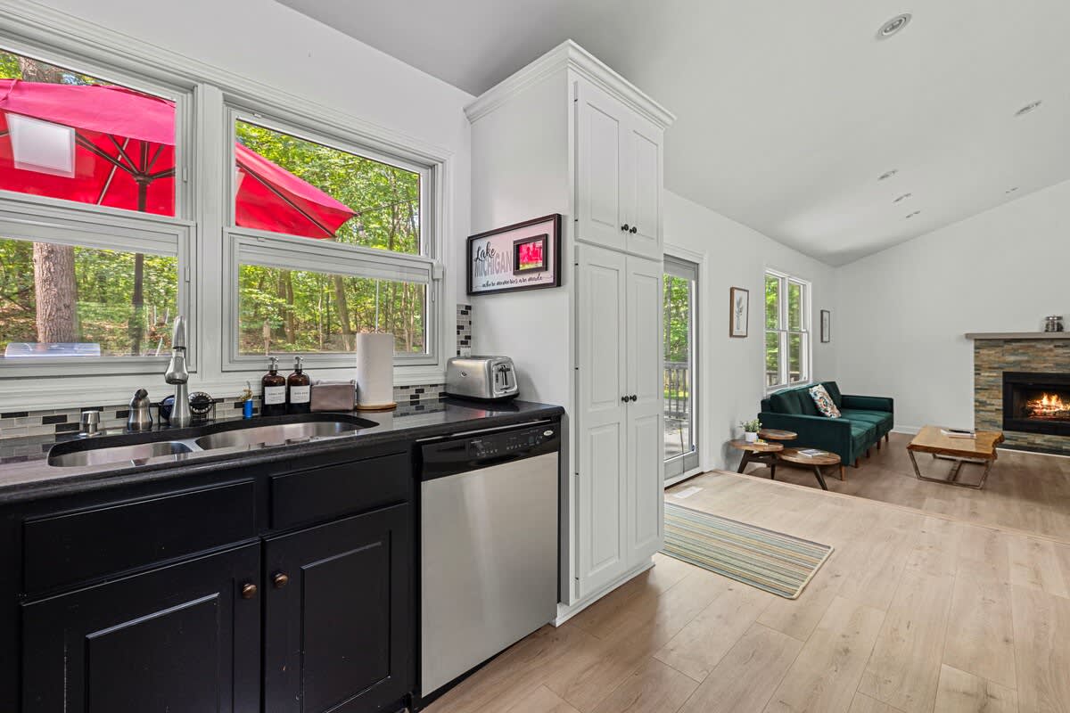 A well-lit kitchen corner featuring a double sink, black countertops, and a mosaic tile backsplash, with large windows overlooking lush trees and a red umbrella, and a view into the living room with a fireplace.