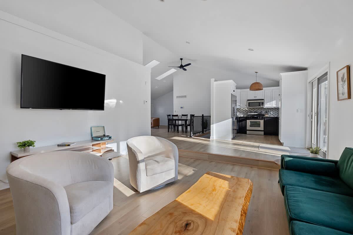 An expansive living room with a large wall-mounted TV, two light-colored swivel armchairs, and a dark green sofa, offering a view into a raised dining area and a black and white kitchen beyond.