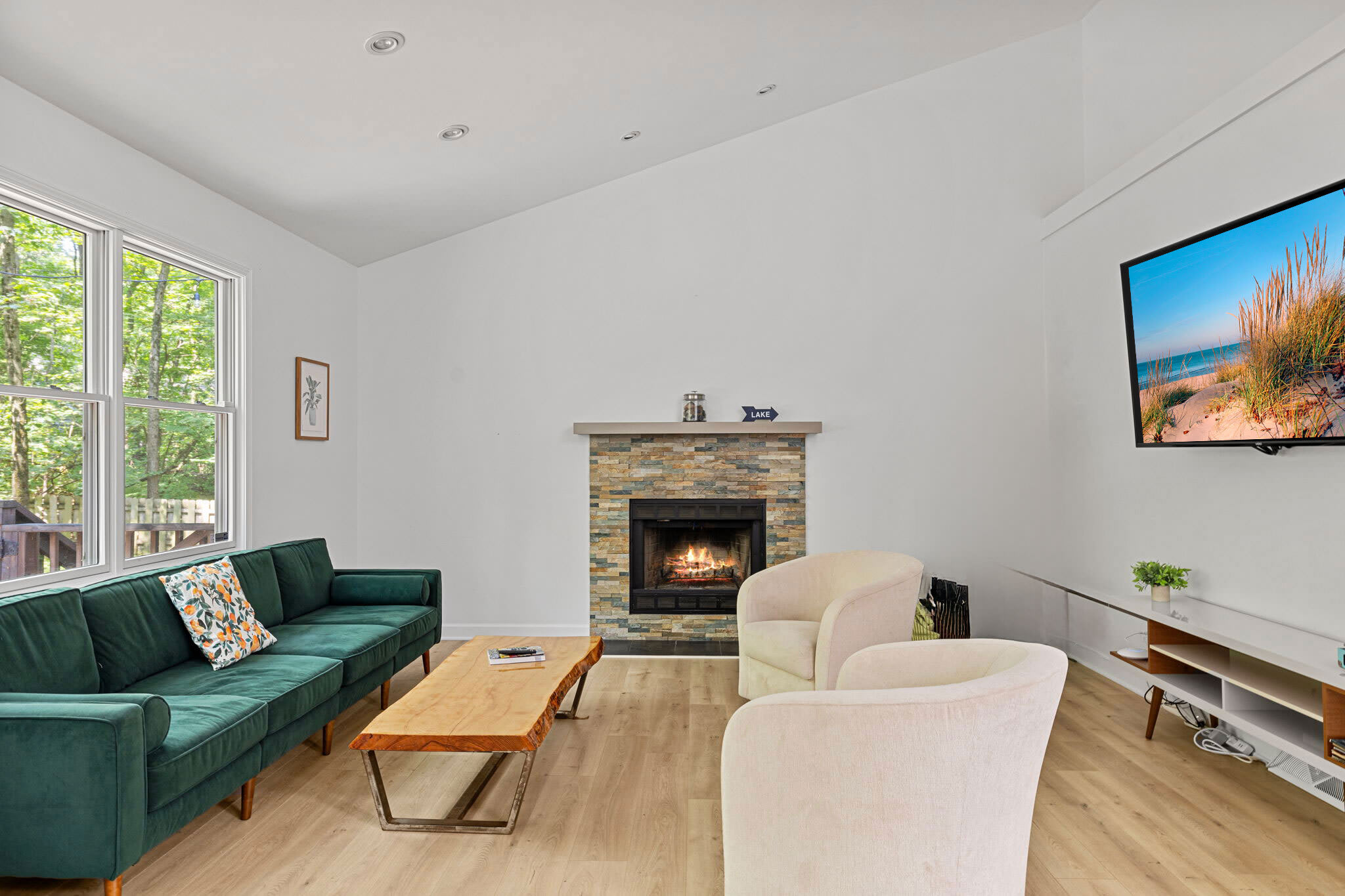 A spacious living room with a dark green velvet sofa, two light-colored armchairs, a stone fireplace, and a large wall-mounted TV displaying a beach scene, all illuminated by natural light from large windows.