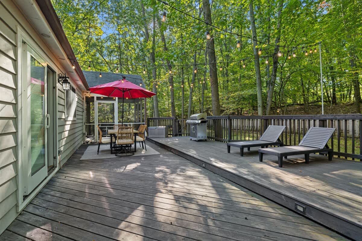 A wide view of the multi-level dark wooden deck, showcasing an outdoor dining area with a red umbrella, a stainless steel grill, and two lounge chairs, all set against a backdrop of dense green woods.