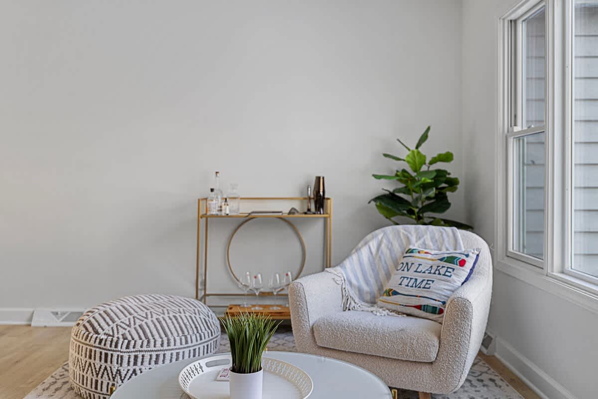 Bright corner of a living room with a white armchair, a patterned pouf ottoman, and a gold bar cart, set against white walls and light wood floors with a large window.