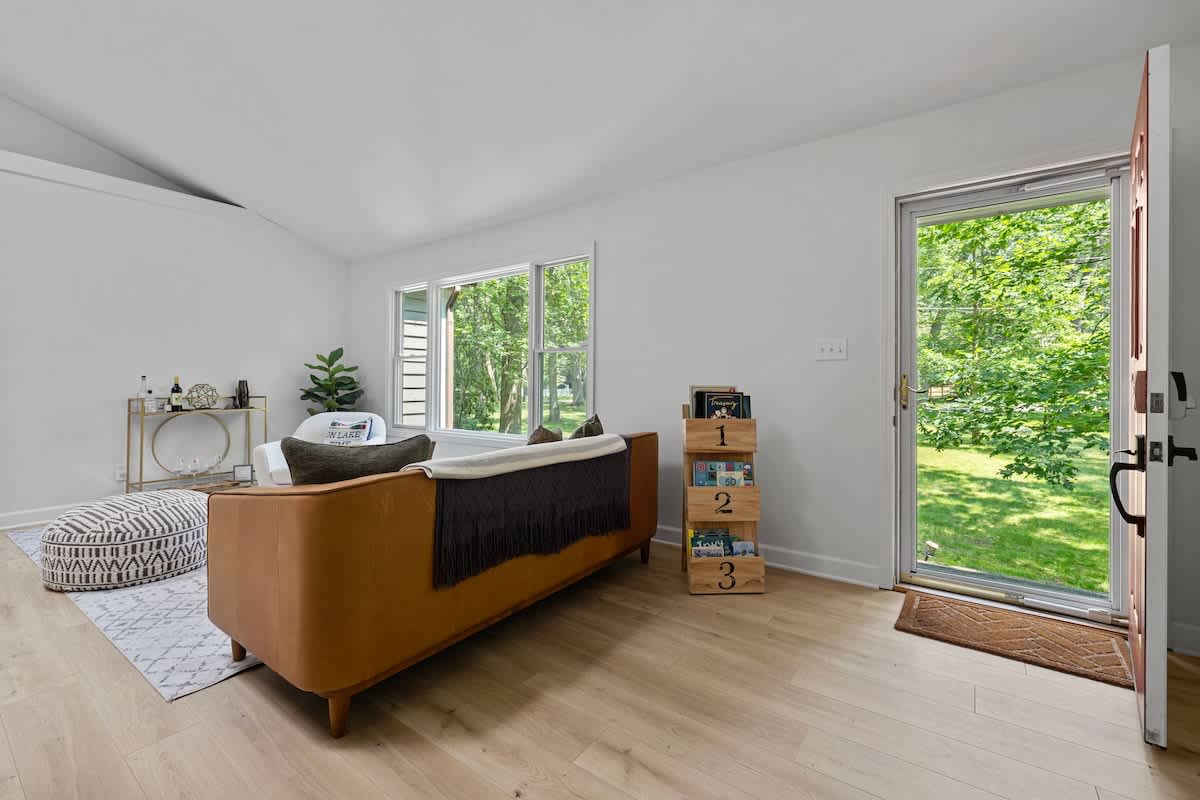 View of the living room from the entrance, showing a brown leather sofa, light wood floors, and a red front door, with a magazine rack and large windows offering a view of the yard.