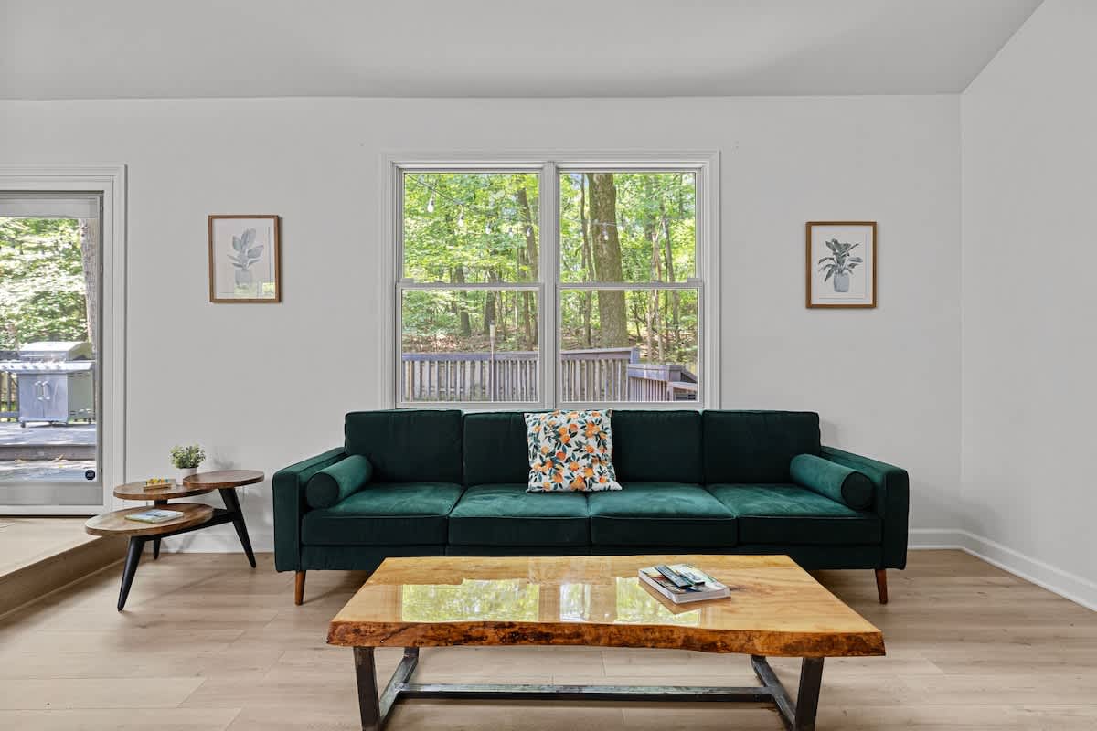 Cozy living area with light wood flooring, a dark green velvet sofa, and a live-edge wood coffee table, featuring a large window overlooking a wooden deck and surrounding trees.