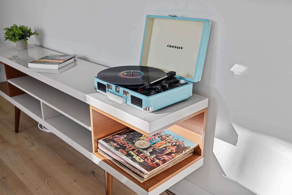 Modern entertainment console with a light blue vintage-style record player and a collection of vinyl records, set against a white wall with light wood flooring.