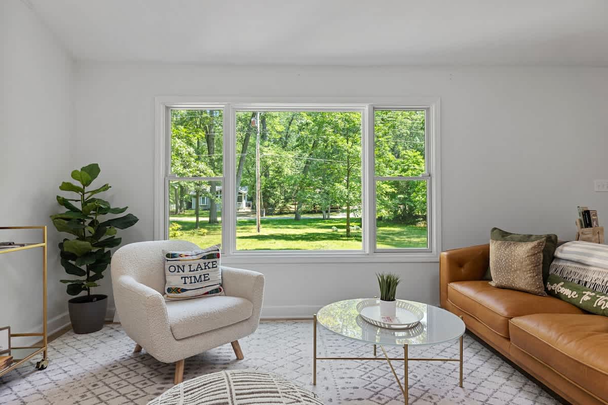 Bright living room with light wood floors, a white shaggy armchair, a brown leather sofa, and a glass coffee table, offering a view of a green yard through large windows.